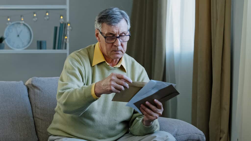 60s elderly man recipient with glasses sorting letters in living room concentrated mature grandfather sitting on sofa looking at correspondence notice bank account caucasian aged pensioner check bills. High quality photo